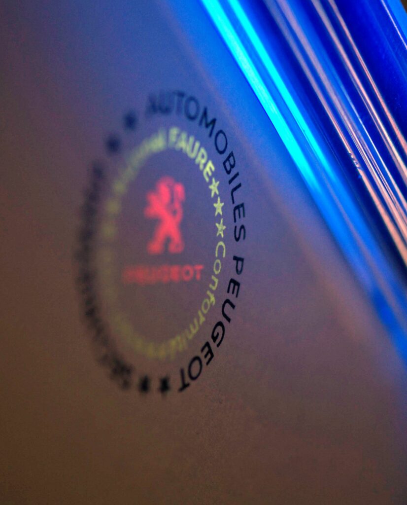 Tampon de sécurité SecuriStamp visible sous lumière ultraviolette (UV). Texte circulaire « SECURI-STAMP * SECURIDOC * » entourant cinq étoiles fluorescentes vertes et un motif central rose fluorescent avec la mention « SECURE A001 ».