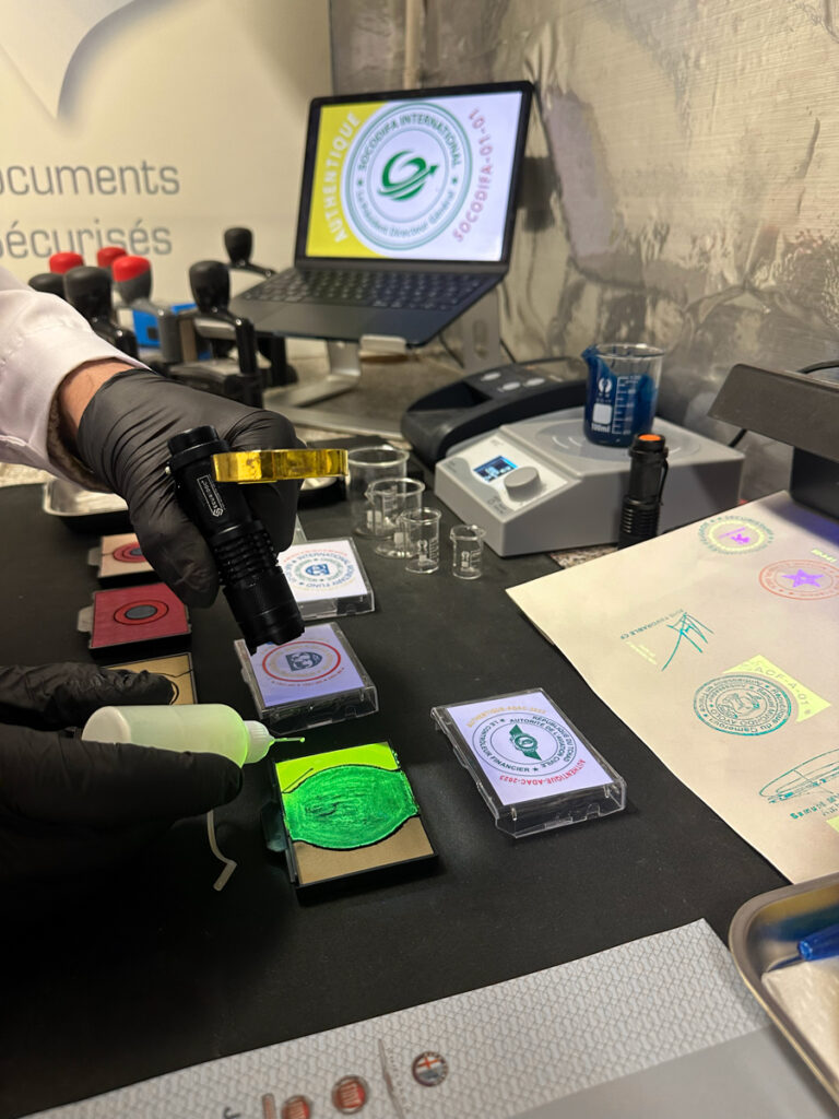 Technicien de laboratoire portant des gants noirs en train d’appliquer une encre fluorescente verte sur un tampon SecuriStamp à l’aide d’une bouteille doseuse, tandis qu’il éclaire le tampon avec une lampe UV. On voit sur le bureau divers tampons, encreurs et documents sécurisés testés.